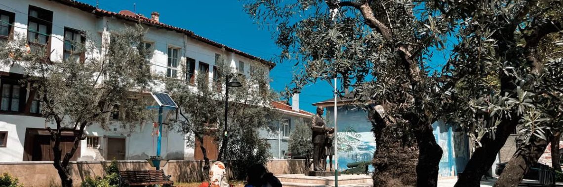 Peaceful village square with benches, olive trees, and historical buildings under clear blue sky.